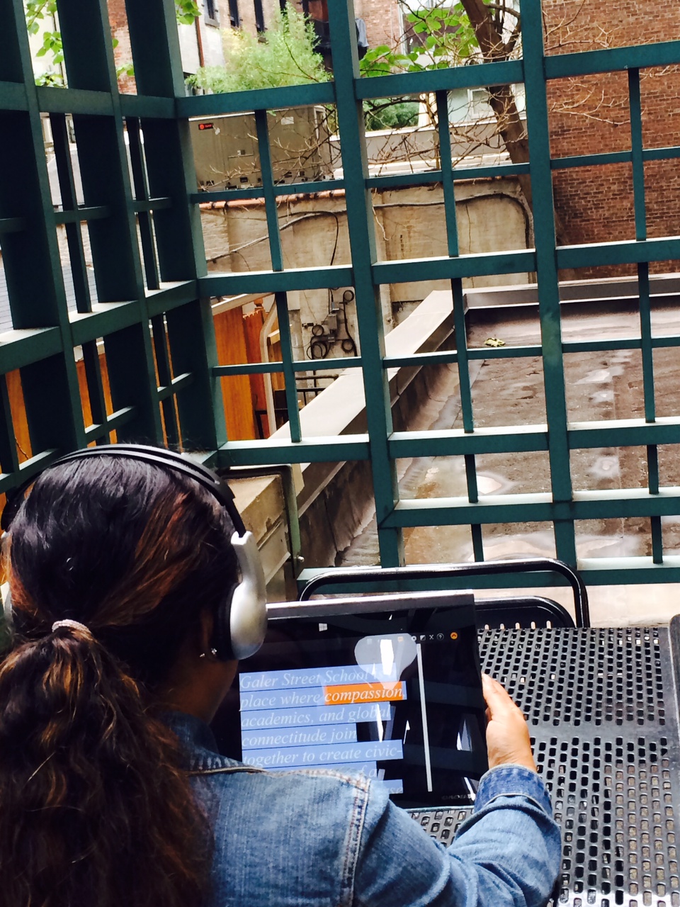 Back of the head of a woman who is wearing headphones and holding a tablet with large words on it, some are highlighted. She sits outside on a terrace and there are trees and buildings in the background. 